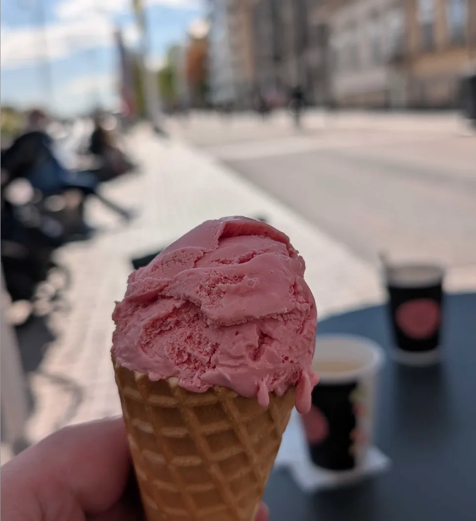 Yksi pallo vaaleanpunaista jäätelöä vohvelissa. Kuvan taustalla Turun Aurajokirantaa sumentuneena. Päivä on aurinkoinen ja kuva huokuu alkavaa kesää.